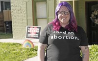 Leah Dean, a native of the Texas Panhandle, poses outside her home Monday, July 3, 2023, in Denver.  In Texas, Dean had been scared to fly an abortion rights banner outside her house. Now in Denver, an LGBTQ+ pride flag flies above the banner in front of her house that proclaims, “Abortion access is a community responsibility.” 
(David Zalubowski / AP Photo)