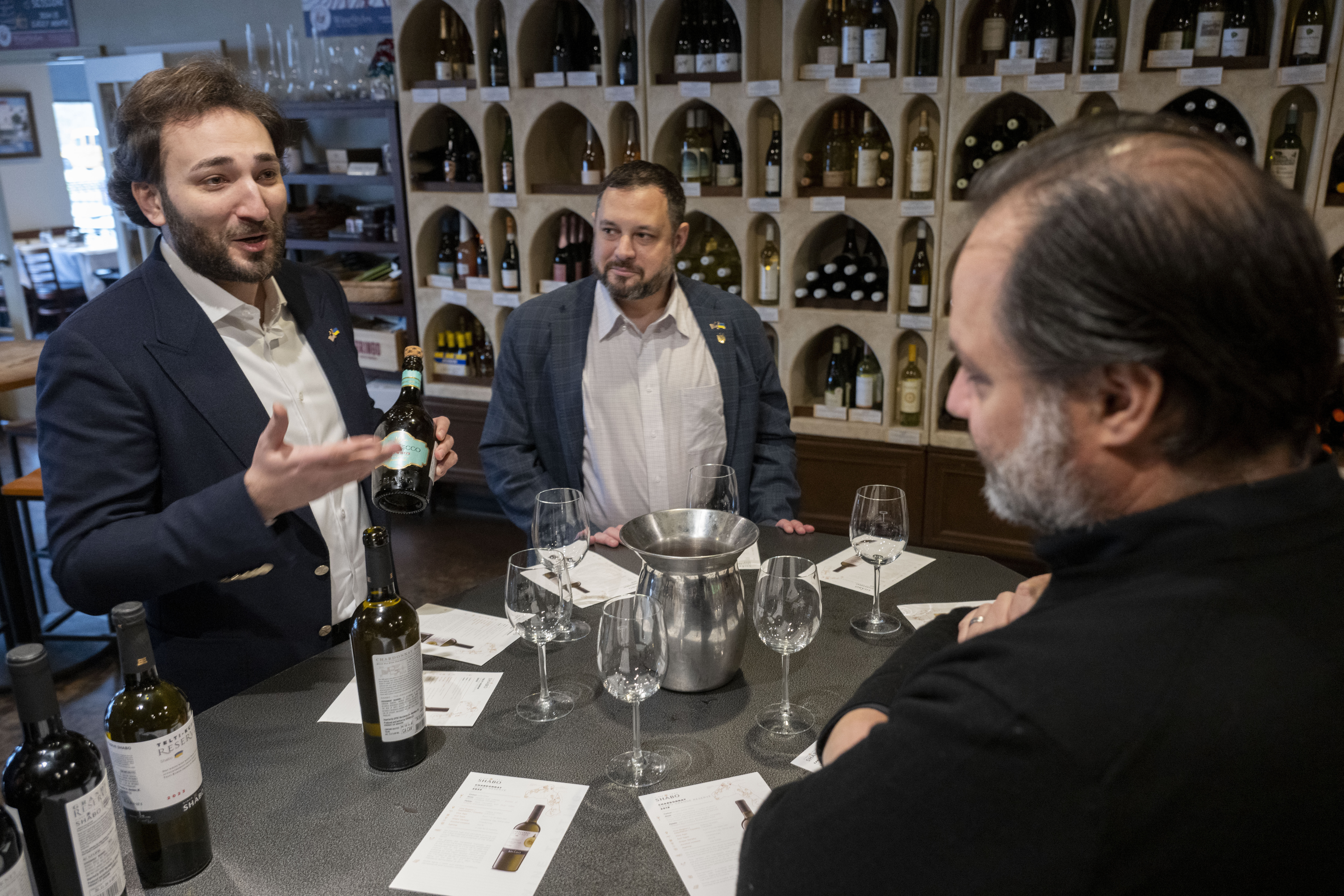 Giorgi Iukuridze (left), owner and CEO of Shabo, a Ukrainian winery, and Sam Lerman (center), CEO of SPYRT Worldwide, introduce Ukrainian wine to the owner of Wine Styles Arthur Lampros (right) at Wine Styles, Dec. 20, 2024 in Dumfries, Va.<br />(Kevin Wolf / AP Photo)