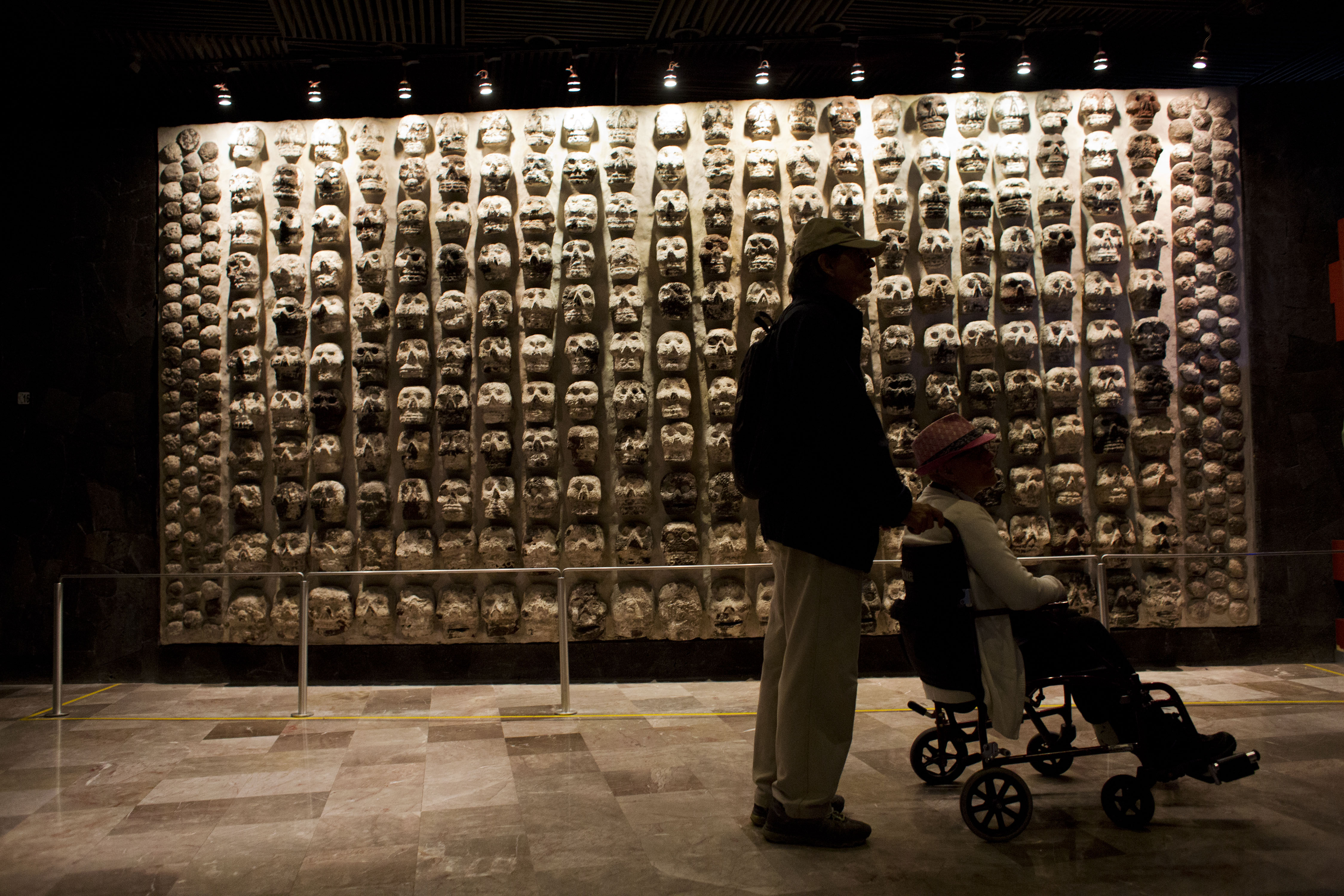 A man pushes a woman in a wheelchair past a wall of ancient stone skulls, excavated at Templo Mayor, that represent sacrificial victims, at the entrance to the Templo Mayor museum in central Mexico City, Friday, Aug. 7, 2015.<br />(Rebecca Blackwell / AP Photo)