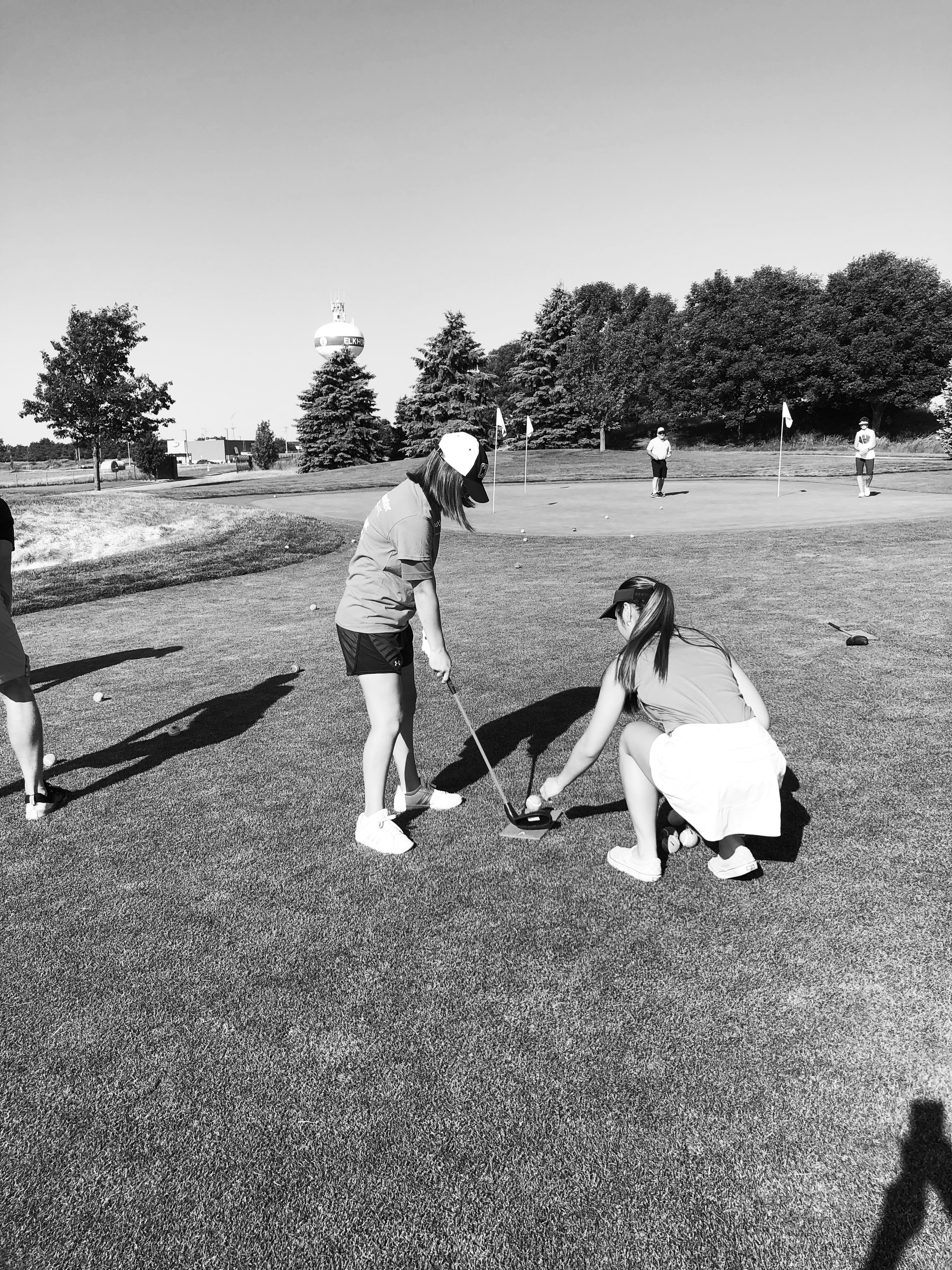 Two young ladies on the golf course. One is visually impaired and holding a golf club. The other is a volunteer golf guide, kneeling down to place the ball near the golf club. (Megan Mackie)