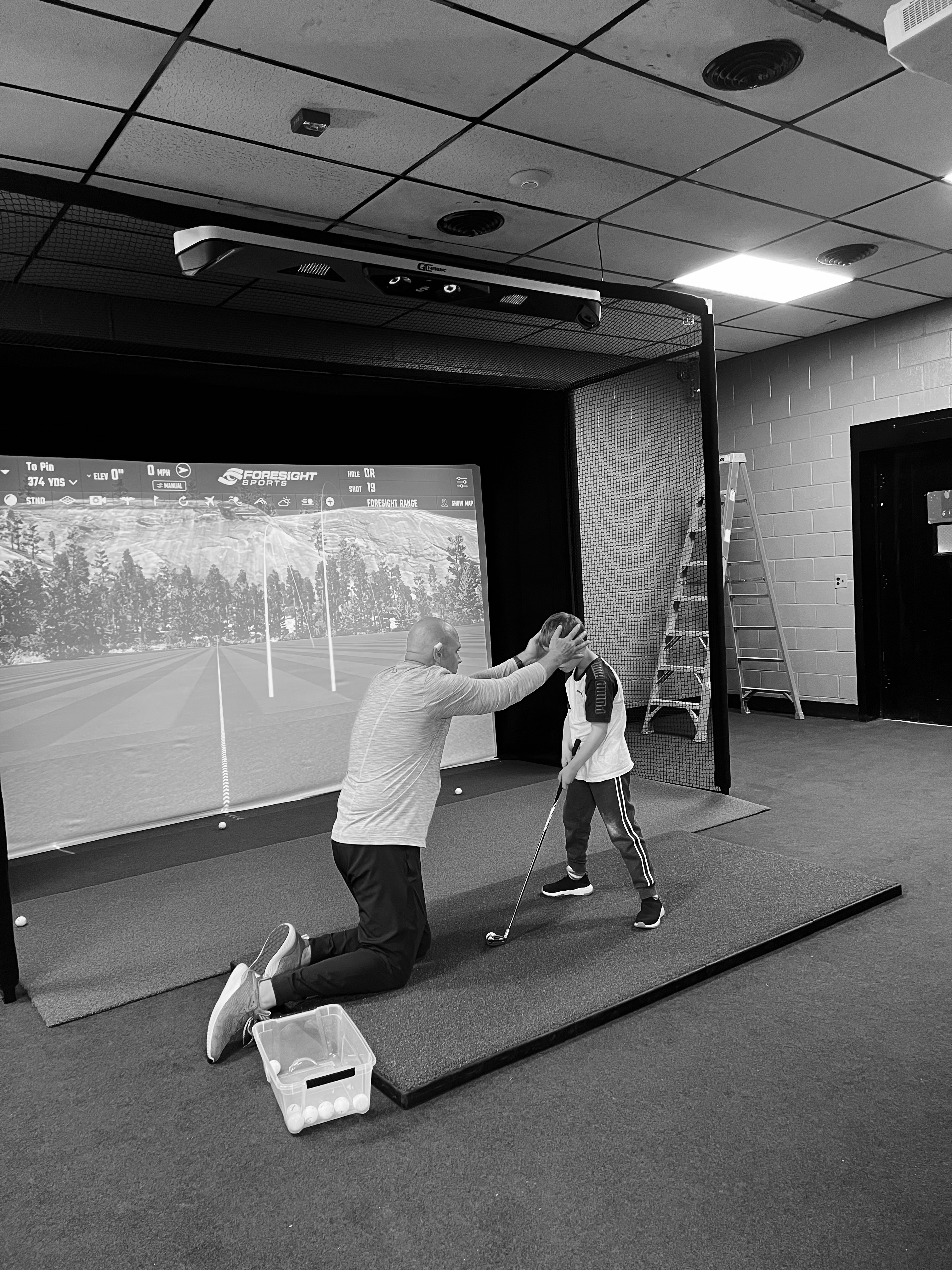 A young boy is holding a golf club. A man is knelt down in front of him. The man has his hands on the boys temples to demonstrate how to line up his body. There is a very large golf simulator screen in the background displaying a picture of a driving range. (Megan Mackie)