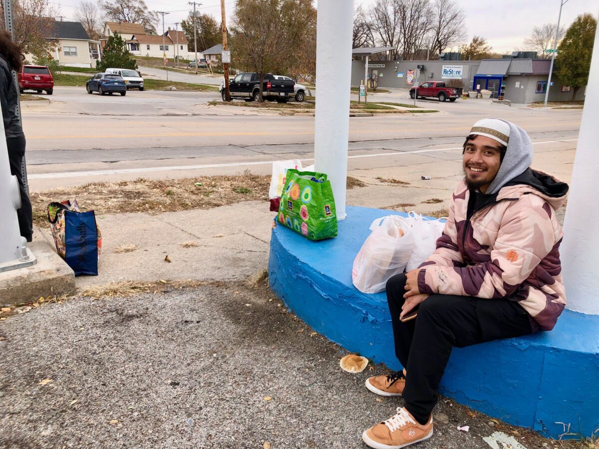 A man who goes by the name of Azitiz waits for the bus after a visit to the food bank. He said he is homeless and sleeping in a storage container on a construction site. “I have all this good food. I’m about to be fed,” he says.<br /> (Cindy Gonzalez / Nebraska Examiner)
