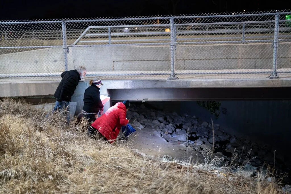 Todd Fleischer (left), Janelle DePuydt (center) and Melissa Neuenfeldt (right) peak under a bridge near the Keystone Trail in Omaha on Jan. 21, 2025. The trio were checking common sleeping spots for Omaha’s unsheltered homeless population as part of the annual nationwide Point-in-Time Count. The survey is required by the U.S. Department of Housing and Urban Development to better understand the nation’s homeless population.<br />(Abioloa Kosoko / Flatwater Free Press)